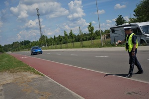 Widok na jezdnię i ścieżkę rowerową. Na jezdni stoi policjant ruchu drogowego zatrzymując do kontroli drogowej nadjeżdżający samochód w niebieskim kolorze