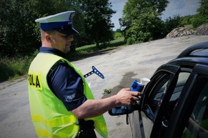 Policjant ruchu drogowego stoi przy zatrzymanym do kontroli czarnym samochodzie trzymając w ręku alkometr