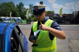 Policjant ruchu drogowego stoi przy zatrzymanym do kontroli niebieskim samochodzie trzymając w ręku terminal płatniczy