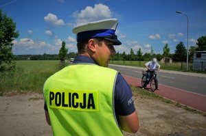 Na pierwszym planie stoi policjant ruchu drogowego w odblaskowej kamizelce. W tle, na ścieżce rowerowej przy jezdni widać mężczyznę na rowerze