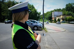 W lewej części zdjęcia na pierwszym planie stoi policjantka z głową skierowaną na widoczne w tle przejście dla pieszych i samochody ustępujące pierwszeństwa pieszemu