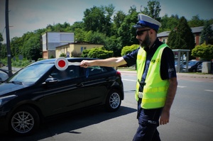 Na pierwszym planie policjant ruchu drogowego idzie trzymając w ręku tarczę STOP, którą wskazuje kierunek jazdy kierowcy czarnego samochodu widocznego na drugim planie