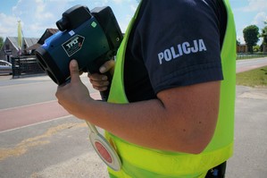 Zbliżenie na ręce policjanta ruchu drogowego w odblaskowej kamizelce, który trzyma tarczę STOP i laserowy miernik prędkości. W tle widać jezdnię