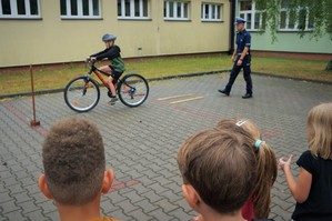 Na placu przed szkołą chłopiec jedzie na rowerze przez tor przeszkód, za chłopcem idzie policjant ruchu drogowego