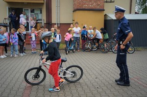 Plac przed szkoła. Na pierwszym planie widać policjanta stojącego za chłopcem na rowerze. Na drugim planie przed budynkiem szkoły stoi grupa dzieci
