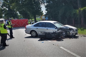 Miejsce wypadku. Po lewej stronie na jezdni stoją policjanci w odblaskowych kamizelkach, którzy pracują na miejscu zdarzenia. Po prawej stronie na jezdni stoją dwa rozbite pojazdy uczestniczące w wypadku. W tle widać czerwony parawan z napisem: STRAŻ