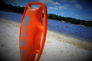 Pomarańczowa boja ratownicza z napisem: WOPR stoi wbita w piasek na plaży, na tle zbiornika wodnego