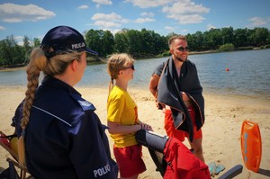 Policjantka stoi z dwojgiem ratowników WOPR na plaży przy zbiorniku wodnym, który widać na drugim planie