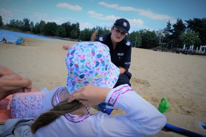 Na plaży na Łysienie policjantka kuca przy dziewczynce w kolorowym kapeluszu, którą widać na pierwszym planie od tyłu. W tle widać zbiornik wodny