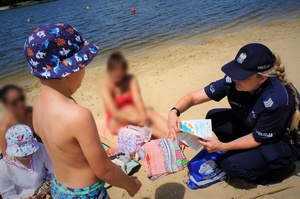 Na plaży nad wodą policjantka kuca przy wypoczywającej rodzinie z dziećmi. Policjantka wręcza małemu chłopcu w kąpielówkach i kapeluszu, ulotkę informacyjną