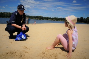 Na plaży policjantka kuca przed małą dziewczynką siedzącą na piasku rozmawiając z nią. W tle widać zbiornik wodny i niebo