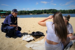 Na plaży policjantka kuca rozmawiając z dwoma nastolatkami odpoczywającymi na kocu. W tle widać zbiornik wodny, las i niebo