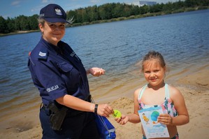 Na plaży, tuż przy brzegu zbiornika wodnego policjantka z małą dziewczynką w stroju kąpielowym pozują do zdjęcia. Policjantka przekazuje dziewczynce odblaskową opaskę. W drugiej ręce dziewczynka trzyma ulotkę informacyjną o bezpieczeństwie nad wodą.