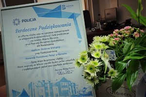 List gratulacyjny od Burmistrza Miasta Bierunia w ramce stoi na stole w gabinecie Komendanta Powiatowego Policji w Bieruniu. Obok listu stoi bukiet kwiatów. W tle widać biurko komendanta