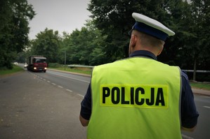 Policjant ruchu drogowego w odblaskowej kamizelce stoi przy jezdni i mierzy prędkość pojazdów