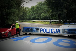 Na pierwszym planie widać listwę sygnalizacyjną radiowozu z napisem POLICJA. Na drugim planie policjant ruchu drogowego stoi przy czerwonym samochodzie zatrzymanym do kontroli. W tle jezdnia, przy której chodnikiem jedzie mężczyzna na rowerze