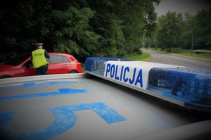 Na pierwszym planie listwa sygnalizacyjna radiowozu, na której widać napis POLICJA. Na drugom planie jezdnia, przy poboczu stoi czerwony samochód zatrzymany do kontroli drogowej przy którym stoi policjant w odblaskowej kamizelce