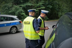 Dwaj policjanci ruchu drogowego stoją przy oknie kierowcy zatrzymanego do kontroli samochodu. W tle widać fragment radiowozu.