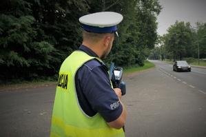 Przy poboczu jezdni stoi policjant ruchu drogowego trzymający miernik prędkości. W kierunku policjanta jezdnią jedzie samochód osobowy w ciemnym kolorze