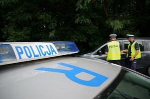 Na pierwszym planie widać fragment dachu radiowozu i listwę sygnalizacyjną z napisem POLICJA. Na drugim planie dwaj policjanci ruchu drogowego stoją przy zatrzymanym do kontroli samochodzie