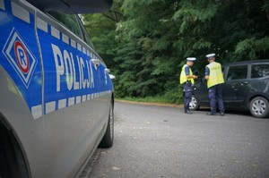 W lewej części zdjęcia zbliżenie na bok radiowozu, na którym widać symbol ruchu drogowego i napis POLICJA. Na drugim planie w prawej części zdjęcia dwaj policjanci ruchu drogowego stoją przy zatrzymanym do kontroli samochodzie