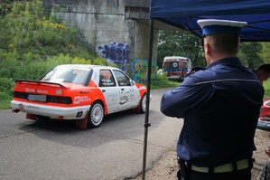 Przy miejscu startowym stoi policjant ruchu drogowego, który przygląda się biało czerwonemu samochodowi wyścigowemu stojącemu na jezdni i oczekującemu na start.