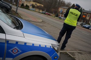Na zdjęciu policjant ruchu drogowego stoi przy jezdni i prowadzi pomiar prędkości pojazdów. Z przodu widać fragment radiowozu