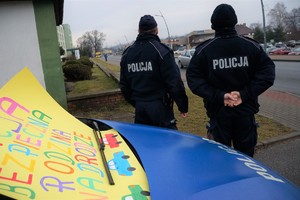 Zdjęcie: z przodu stoi radiowóz, na którym jest plakat akcji. Z tyłu stoją dwaj dzielnicowi przyglądający się jezdni